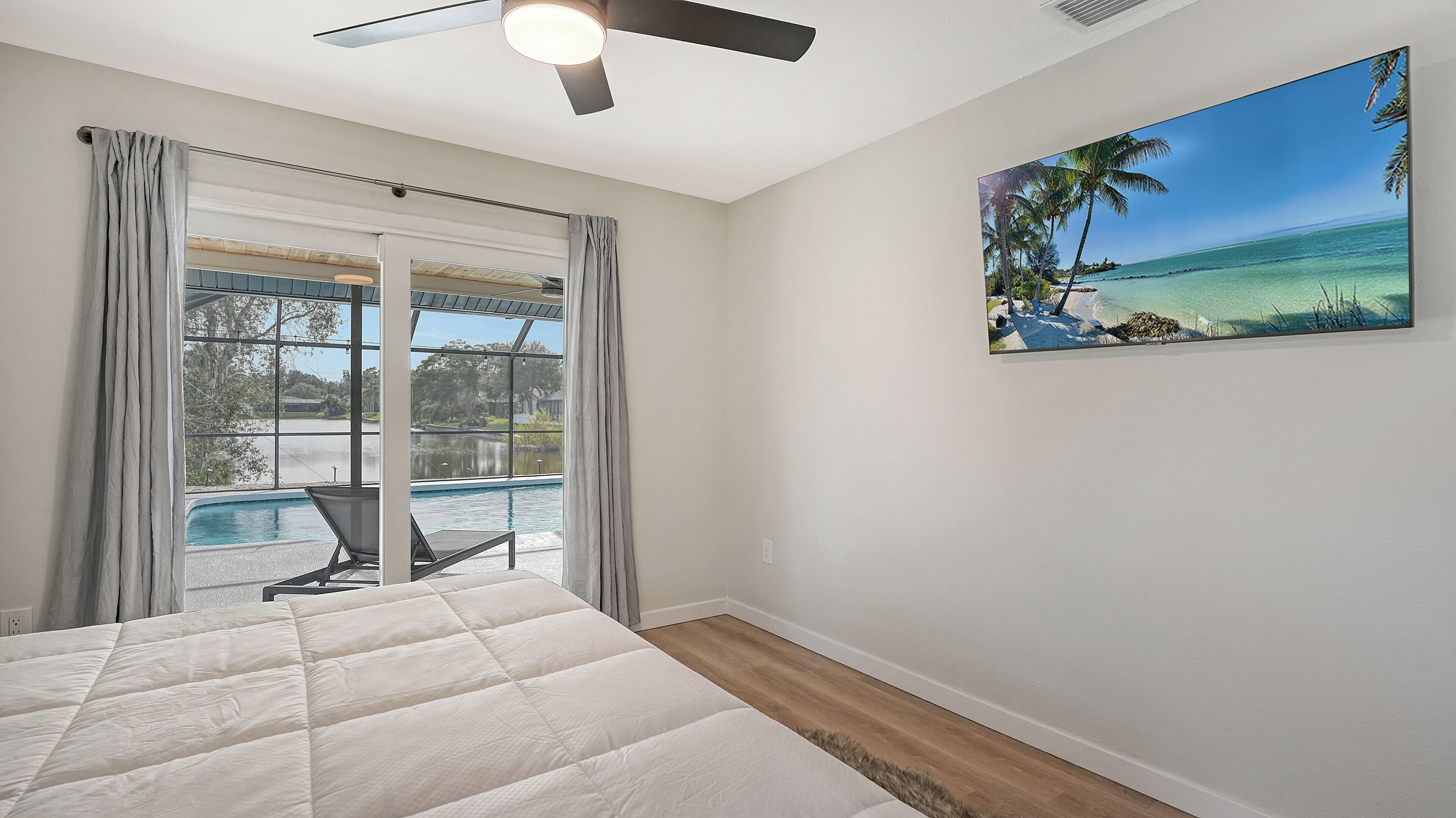 Une chambre avec un lit, un ventilateur de plafond, un tableau représentant une plage, et une vue sur une piscine et un espace extérieur.