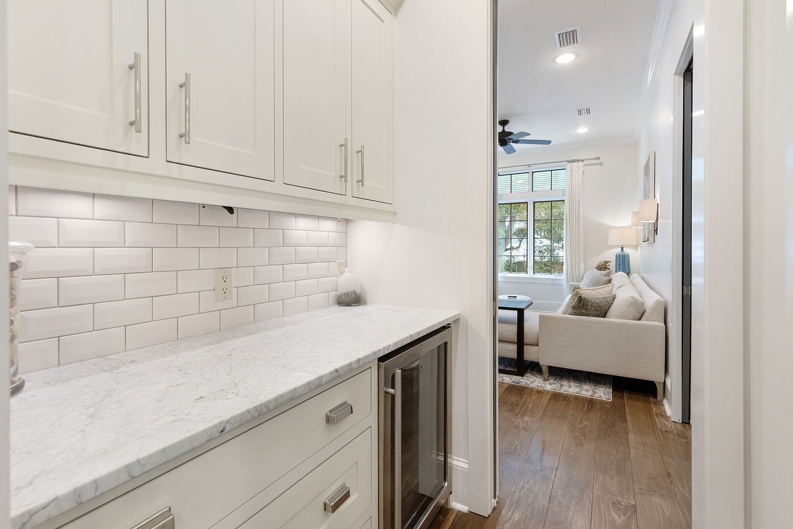 A modern kitchen with white cabinets, a marble countertop, and a view into a living area with a sofa and a table.