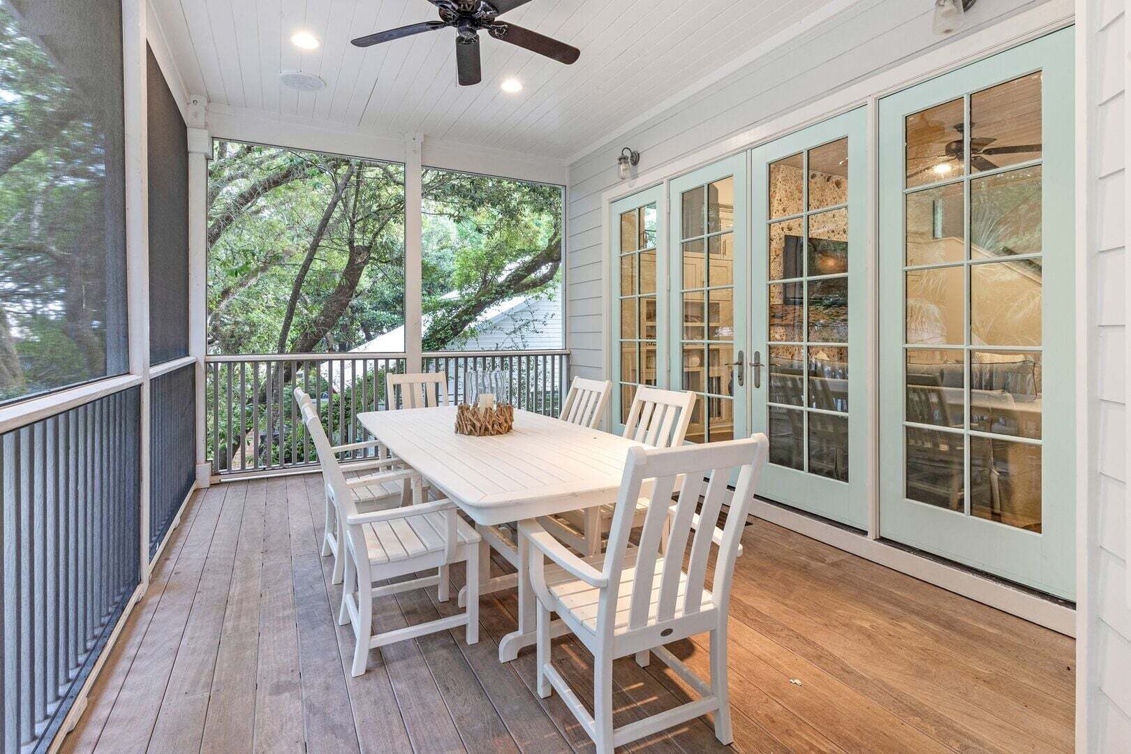 Un espace extérieur abrité, aménagé pour les repas, avec une table et des chaises, un ventilateur de plafond et de grandes fenêtres offrant une vue sur les arbres.