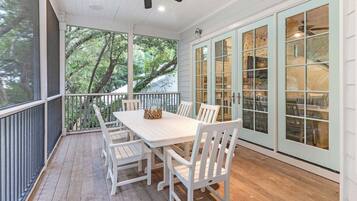 Un espace extérieur abrité, aménagé pour les repas, avec une table et des chaises, un ventilateur de plafond et de grandes fenêtres offrant une vue sur les arbres.