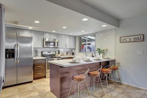A modern kitchen with stainless steel appliances, a central island with bar stools, and a 'let's eat' sign on the wall. - Osborn (Scottsdale)