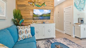 A living room with a blue sofa, a patterned throw pillow, a wooden wall with a mounted TV, a white cabinet, and a kitchen area with a coffee machine. - Free Bird @ The Oasis | Relaxed Coastal Cottage Retreat (Rockport)