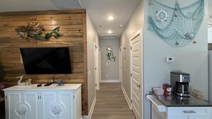 A modern kitchen with a coffee maker, a TV mounted on a wooden wall, and a decorative wall hanging. - Free Bird @ The Oasis | Relaxed Coastal Cottage Retreat (Rockport)