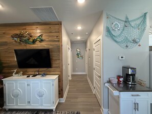 A modern kitchen with a coffee maker, a TV mounted on a wooden wall, and a decorative wall hanging. - Free Bird @ The Oasis | Relaxed Coastal Cottage Retreat (Rockport)