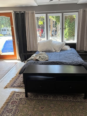 A bedroom with a bed, a nightstand, a window with curtains, and a view of the outside.
