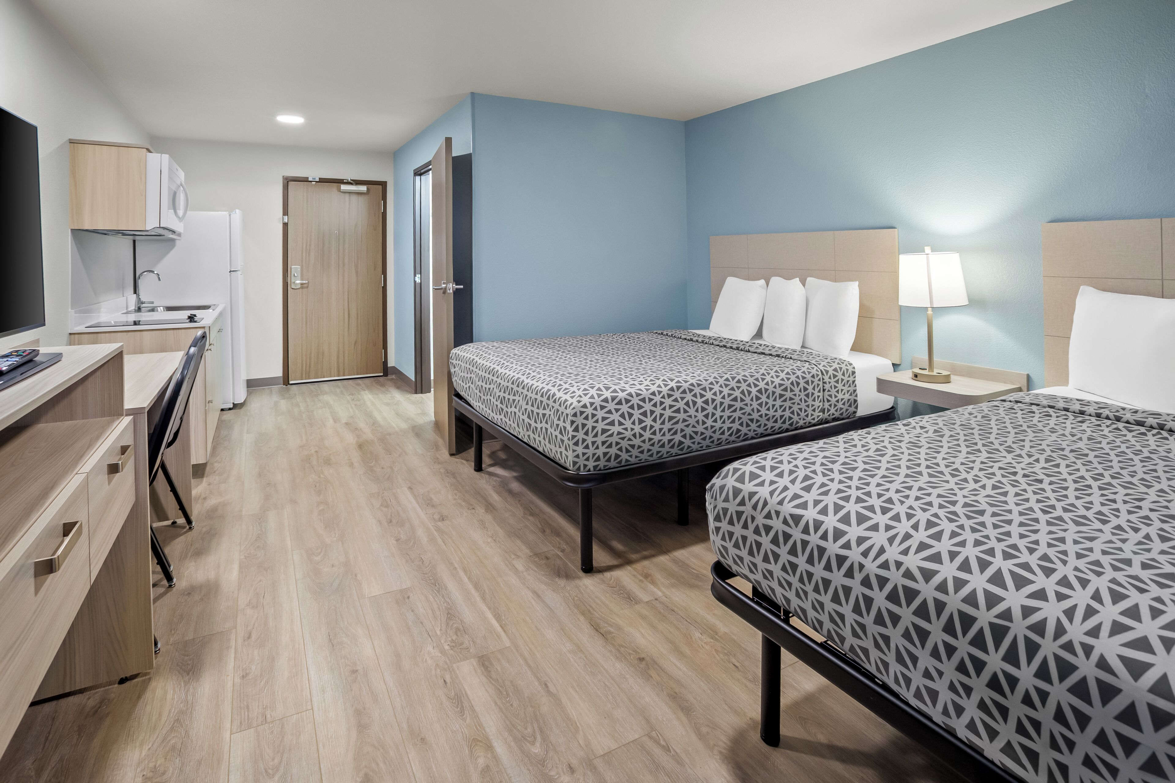A hotel room with two beds, a desk, a kitchenette, and a TV.