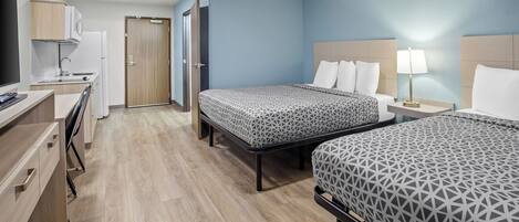 A hotel room with two beds, a desk, a kitchenette, and a TV.
