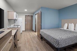 Une chambre comprenant un lit, un bureau, un coin cuisine et une porte donnant sur une autre pièce.