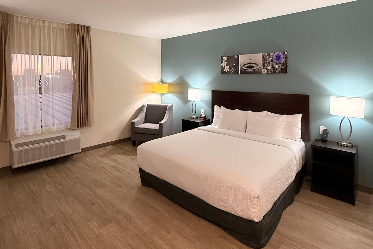 Une chambre d’hôtel avec un grand lit, deux tables de chevet avec des lampes, une chaise, une fenêtre avec des rideaux et un mur orné de trois tableaux encadrés.