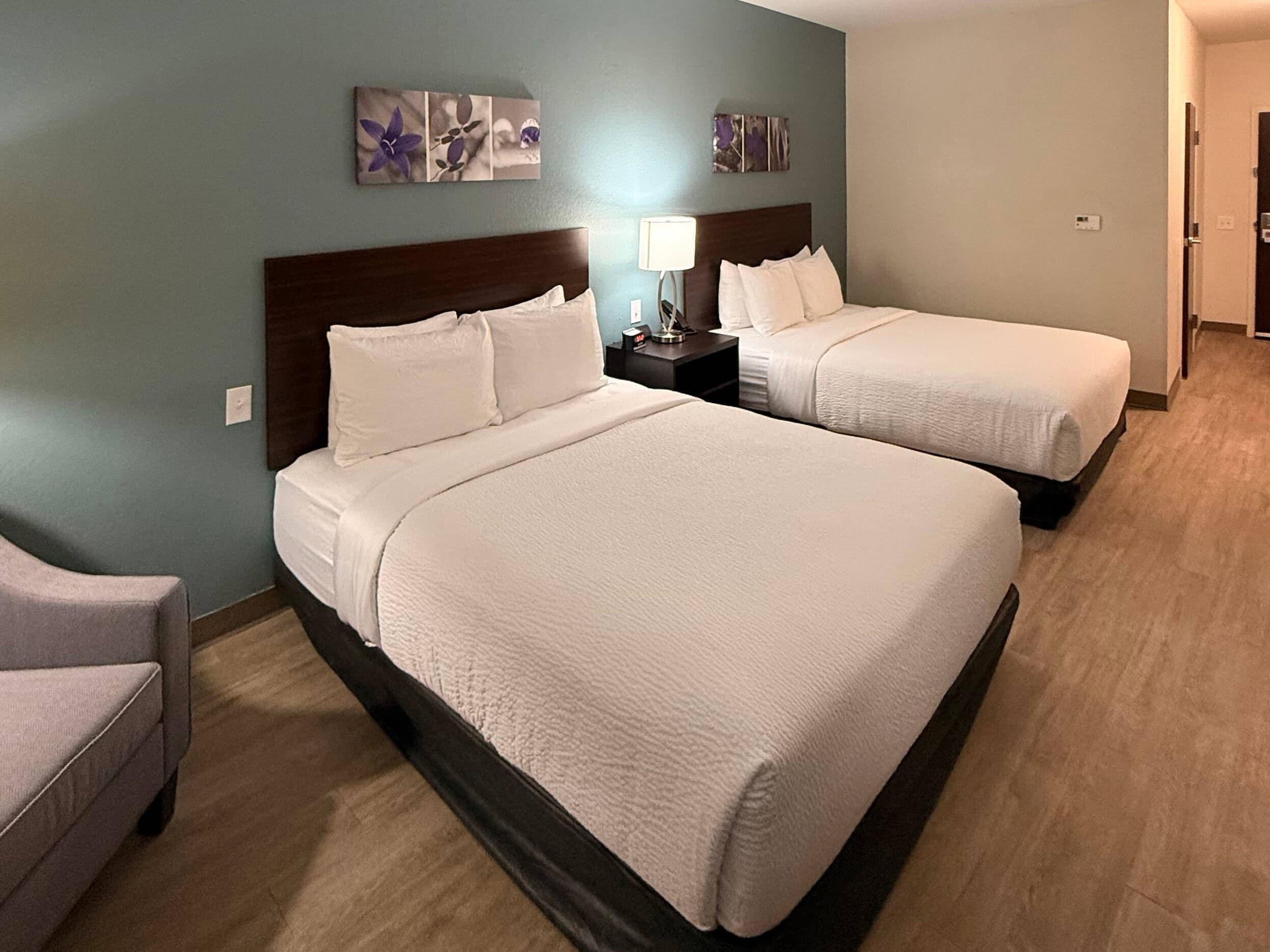 A hotel room with two beds, a nightstand with a lamp, and a chair.