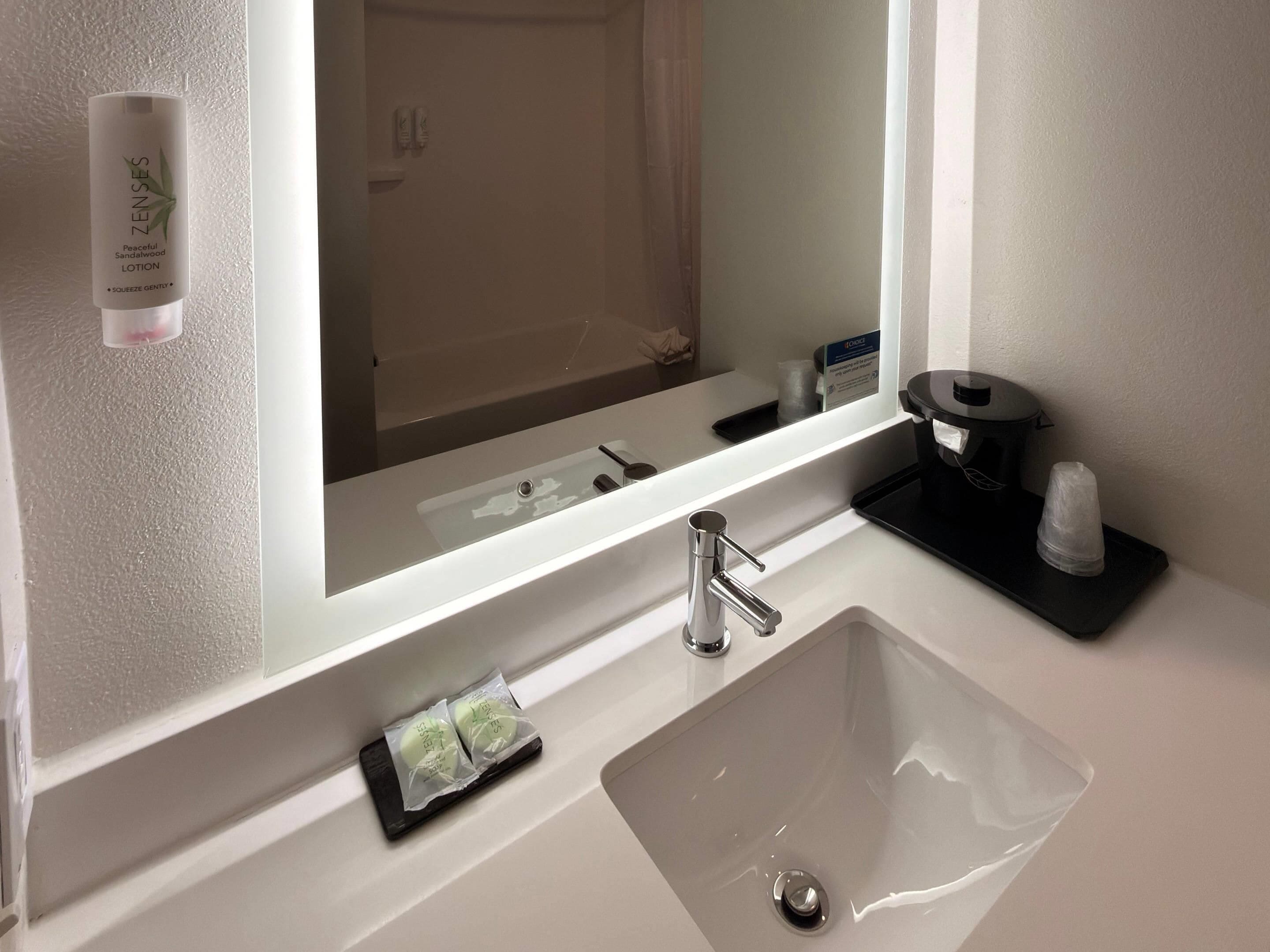 Une salle de bain moderne avec un grand miroir, un lavabo blanc, un porte-savon noir et un flacon de lotion fixé au mur.