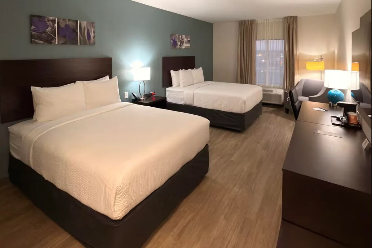 A hotel room with two beds, a desk, a chair, a lamp, and artwork on the walls.