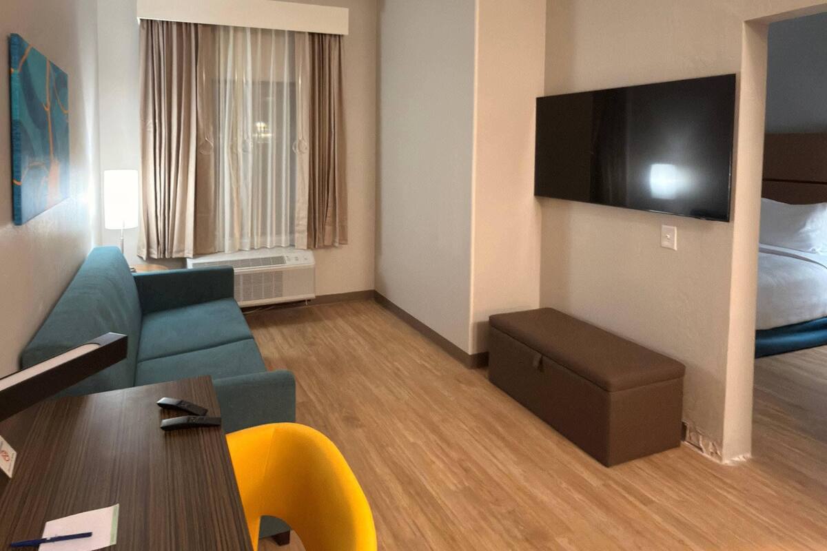 Une chambre d’hôtel avec un plancher en bois, un canapé, une table à manger, un banc, un téléviseur fixé au mur et une fenêtre avec des rideaux.