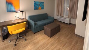 A modern living room with a wooden floor, a blue sofa, a yellow office chair, a brown ottoman, a desk with a lamp, and two abstract paintings on the wall. - Mainstay Suites - Oklahoma City Airport (Oklahoma City)
