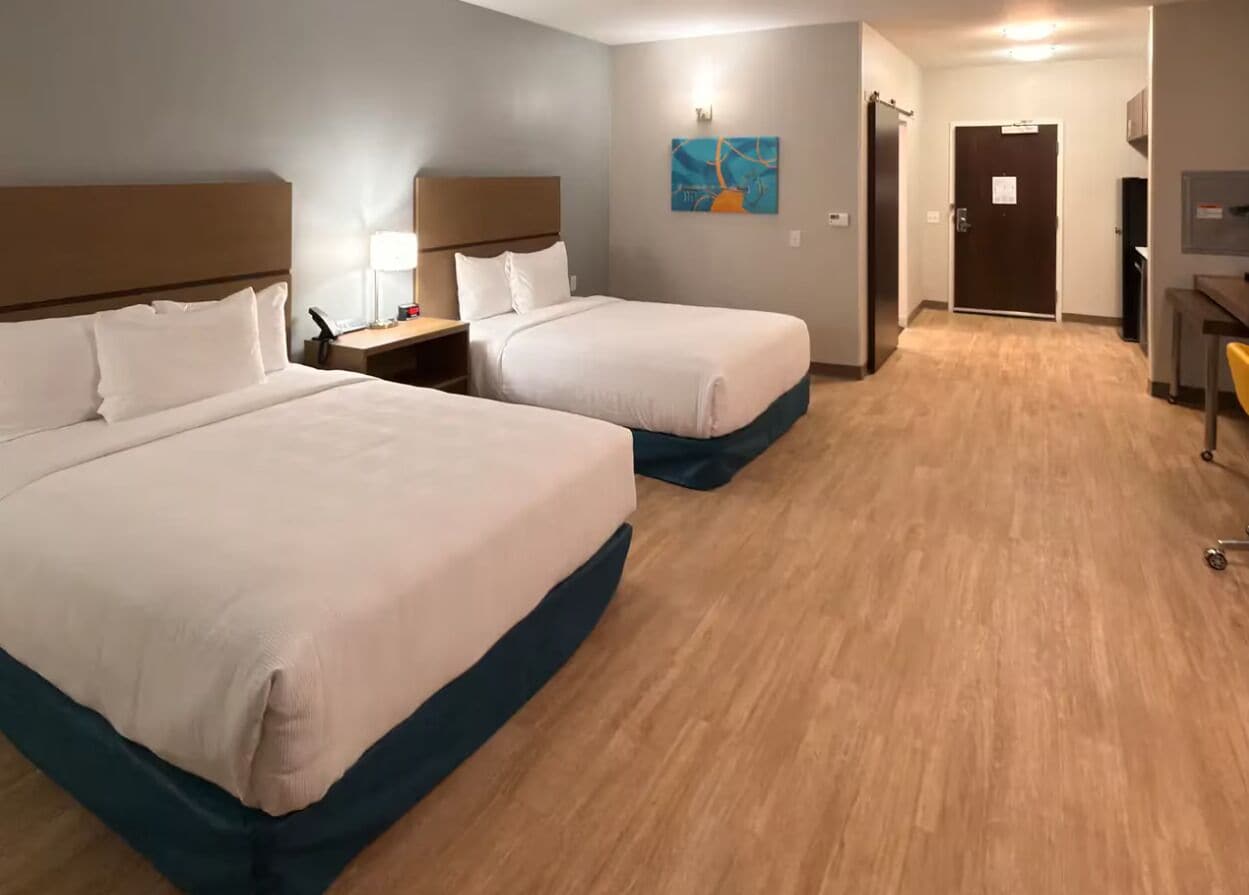 A hotel room with two beds, a desk, a chair, a lamp, a painting, and a door.