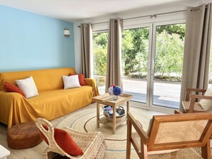 Ein Zimmer mit einem gelben Sofa, einem Korbstuhl, einem Holztisch mit einer Vase und Blick auf Grünpflanzen durch große Fenster.
