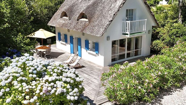 Een huis met rieten dak, een balkon en een houten terras met buitenmeubilair.