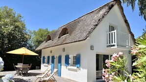 Een wit gebouw met rieten dak, een balkon en een houten terras met lounge stoelen en een parasol.