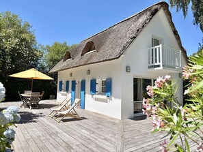 Ein weißes Gebäude mit Strohdach, einem Balkon und einer Holzterrasse mit Liegestühlen und Sonnenschirm.