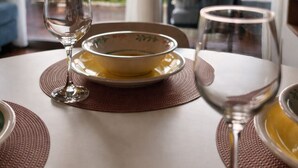 Een eettafel met een schaal en wijnglazen, gedekt met bruine placematen, in een kamer met grote ramen en uitzicht op groen.