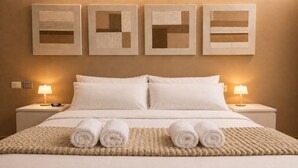 Een net opgemaakt bed met opgerolde handdoeken, een gebreid plaid en vier ingelijste kunstwerken aan de muur.