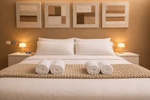 Ein ordentlich bezogenes Bett mit aufgerollten Handtüchern, einer gemusterten Decke und vier gerahmten Kunstwerken an der Wand.