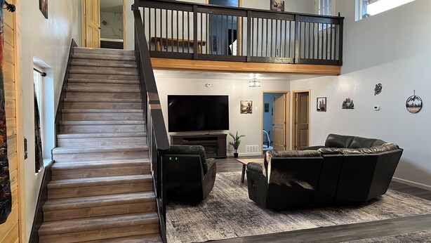 En rummelig opholdsstue med en trætrappe, stort fladskærms-tv, lædersofa og et højt loft med synlige bjælker.