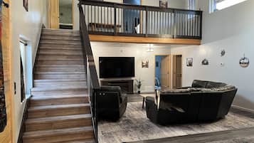 Un vaste espace de vie agrémenté d’un escalier en bois, d’un grand téléviseur à écran plat, d’un canapé en cuir et d’un plafond haut avec des poutres apparentes.