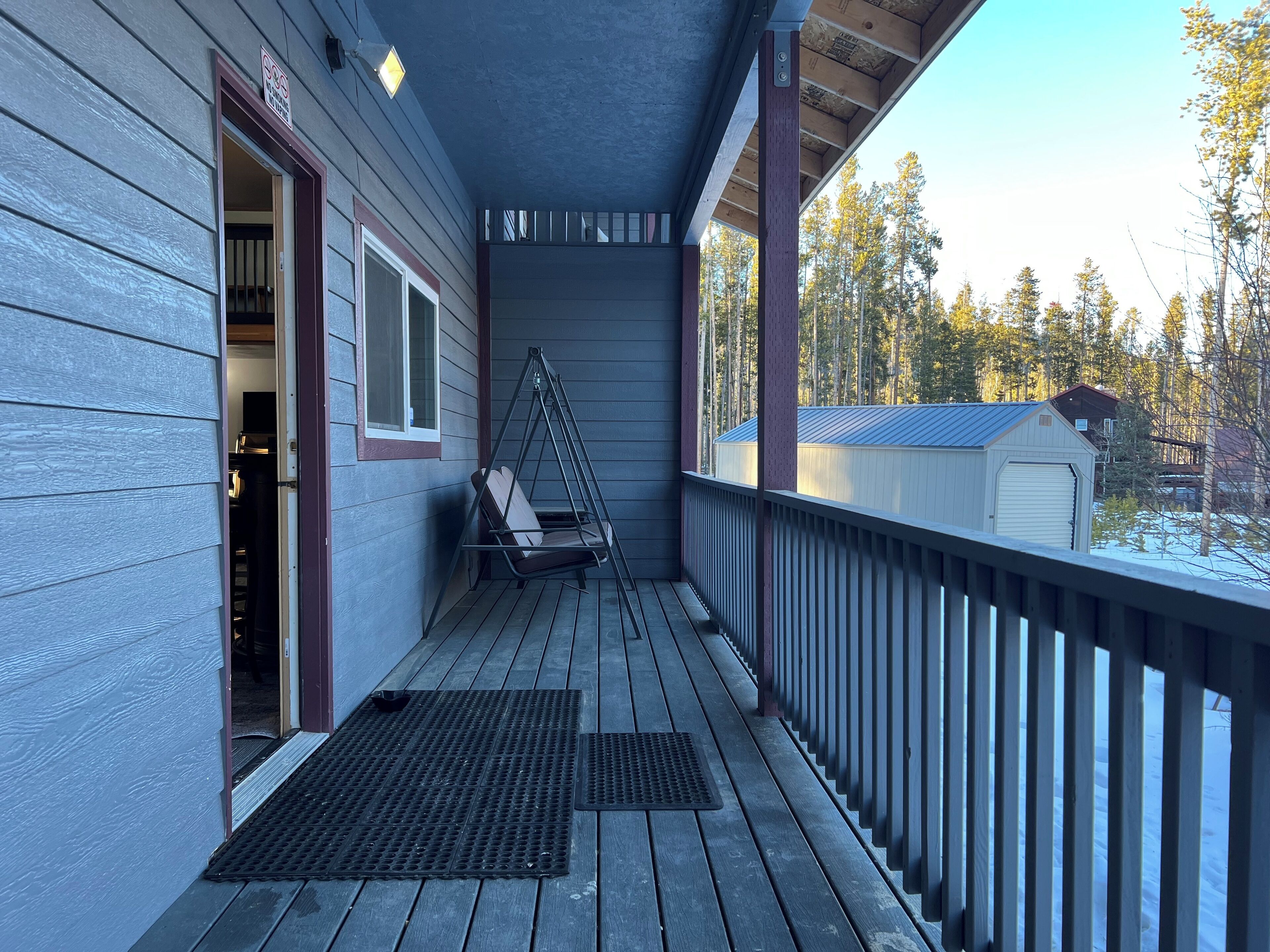 Une véranda en bois avec une chaise suspendue, un espace abrité et une vue sur un paysage enneigé parsemé d’arbres.