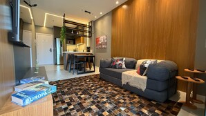 Ein modernes Wohnzimmer mit Schachbrettmuster-Boden, einem grauen Sofa mit Zierkissen, einem kleinen Tisch mit Büchern und einer Küchenzeile im Hintergrund.