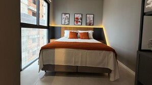Ein modernes Schlafzimmer mit großem Fenster, einem Bett mit weißer und orangefarbener Bettwäsche sowie drei gerahmten Bildern an der Wand.