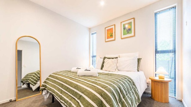 A hotel room with a bed, a bedside table with a lamp, a mirror, and two framed pictures on the wall. - Sydenham | Patio + Free Parking Near CBD (Christchurch)