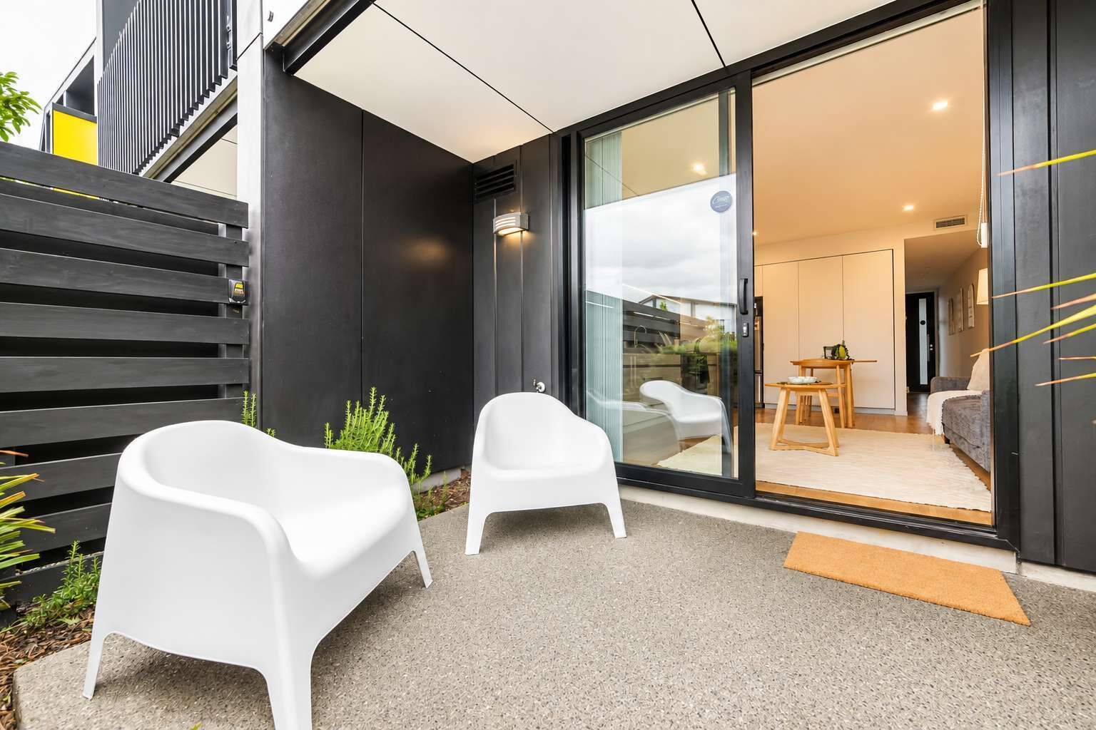 A modern exterior with a sliding glass door leading to a living area, two white chairs outside, and a small outdoor table.