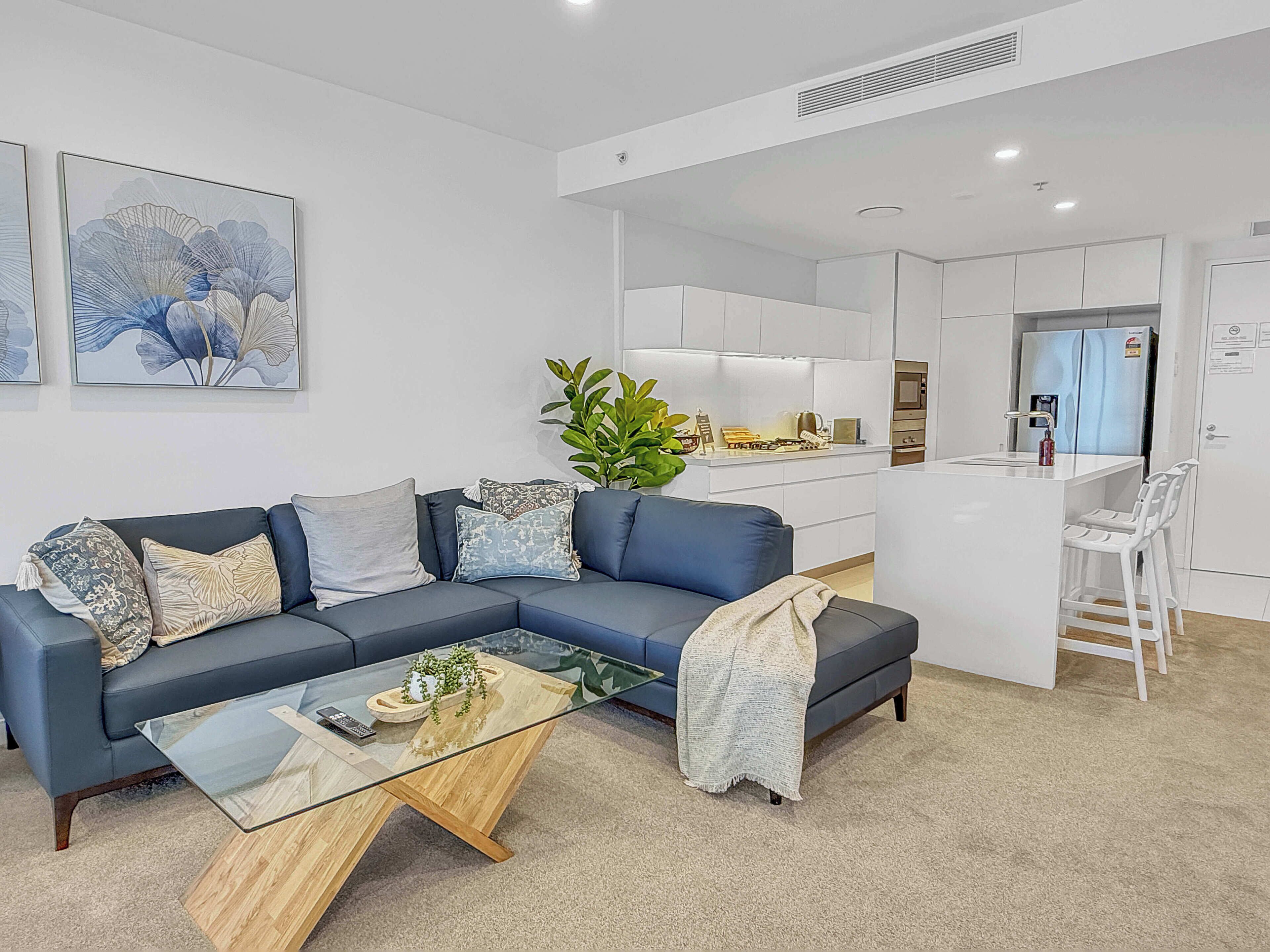 A modern living room with a blue sofa, a glass coffee table, and a kitchen area in the background.