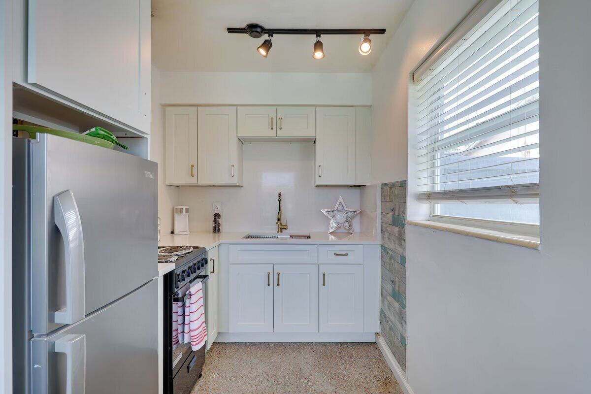 A compact kitchen with white cabinets, a stainless steel refrigerator, a stove, and a window with blinds.