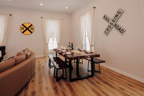A dining area with a wooden table set for four, a leather sofa, and a window with curtains. - Mancelona Train Depot - A Northern Michigan Gem - 15 minutes from Boyne Mountain (Mancelona)