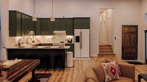 Una cocina moderna con armarios verde oscuro, encimera blanca y una isla central con taburetes. Se observa una mesa de comedor de madera con velas y un banco. Hay un refrigerador y un horno en la cocina.