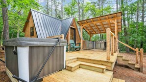 A cabin with a hot tub, wooden deck, and a covered outdoor seating area in a forest.