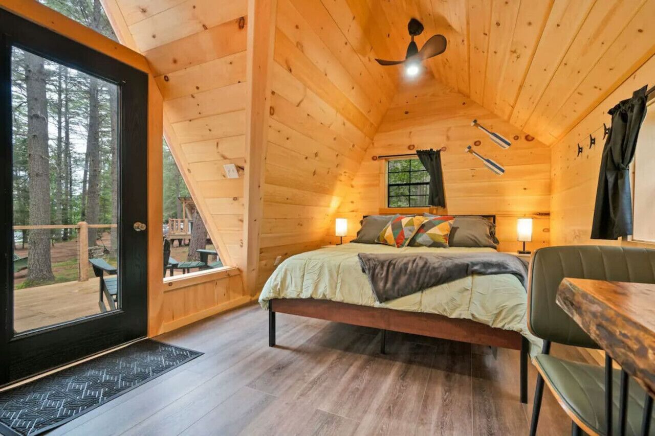 A cozy wooden cabin bedroom with a bed, bedside lamps, and a view of the forest through a glass door.