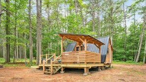 A wooden cabin with a sloped roof and a covered deck, surrounded by a forest.