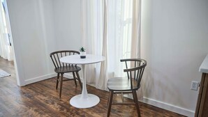 Un petit coin repas avec une table ronde blanche et deux chaises en bois, une plante en pot posée sur la table et un rideau blanc en arrière-plan.