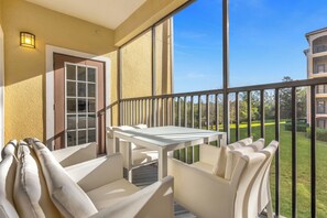 A balcony with a white table and chairs, a door, and a view of a well-maintained lawn and building in the background. - 6105 (Orlando)