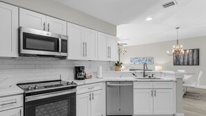 A modern kitchen with white cabinetry, a stainless steel microwave, a built-in oven, a dishwasher, and a central island with a sink and a chandelier. - 6105 (Orlando)
