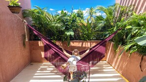 Une personne se détend dans un hamac, dans une cour ensoleillée, entourée d’une végétation tropicale luxuriante.