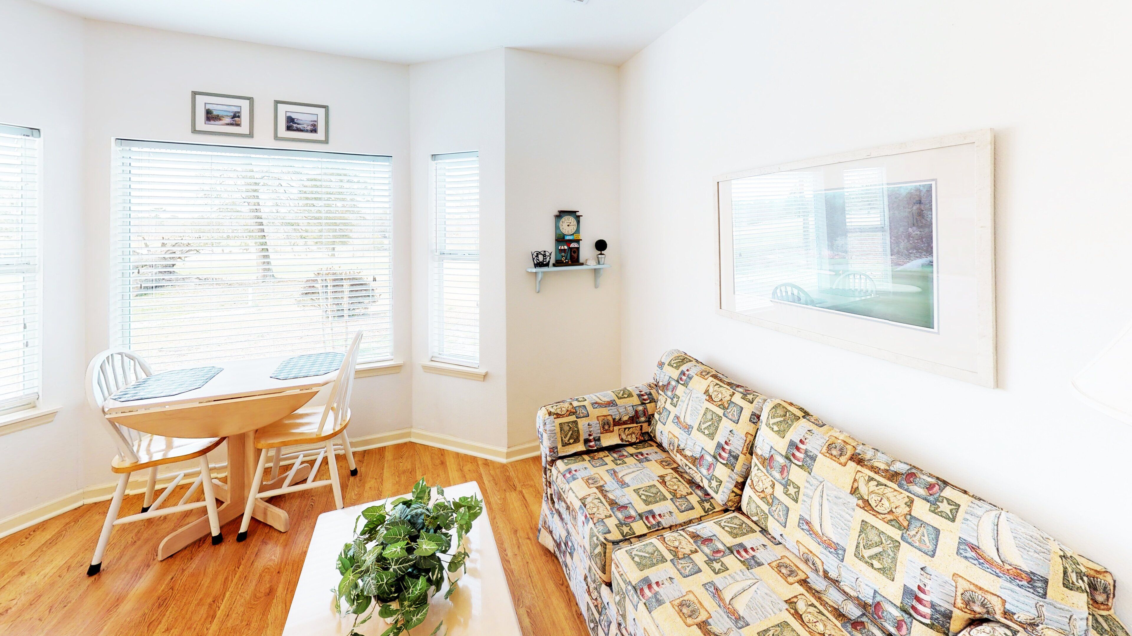 A living room with a wooden dining table, two chairs, a patterned sofa, a coffee table with a plant, and large windows with blinds.