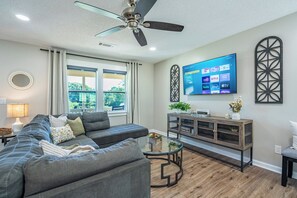 Ein Wohnzimmer mit grauem Sofa, einem Glastisch, einem Fernseher an der Wand und einem Deckenventilator.