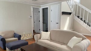 Una sala de estar con un sofá color crema, un sillón azul, un reposapiés floral y una mesa de centro de madera. Se observa una escalera con barandillas blancas y una puerta que da a otra habitación.