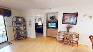 A living room with a TV, a bookshelf, a dining area with chairs, and a door leading to another room. - River Creek 1 Unit 401 (Sunset Beach)
