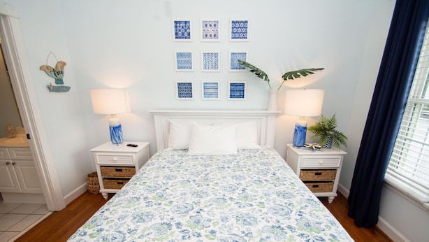 A bedroom with a floral bedspread, two bedside tables with lamps, a white headboard, and a wall with framed artwork. - River Walk 304 (Myrtle Beach)