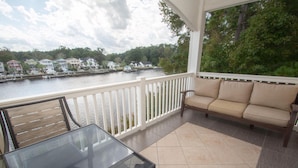 A covered balcony with a glass table and a beige sofa, overlooking a body of water with houses and trees in the background. - River Walk 304 (Myrtle Beach)